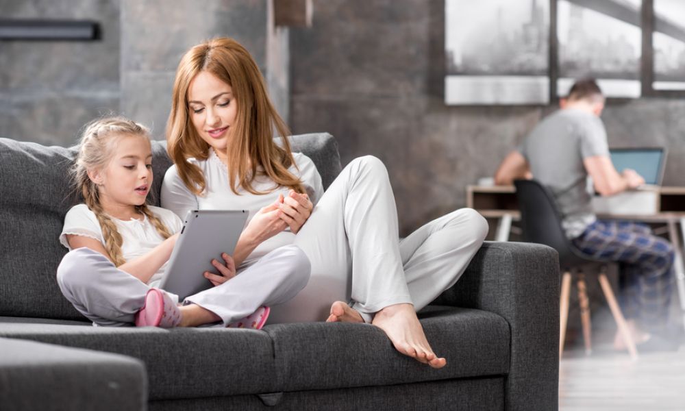 a woman and child sit on a couch while looking at a tablet. A man sits in the background.