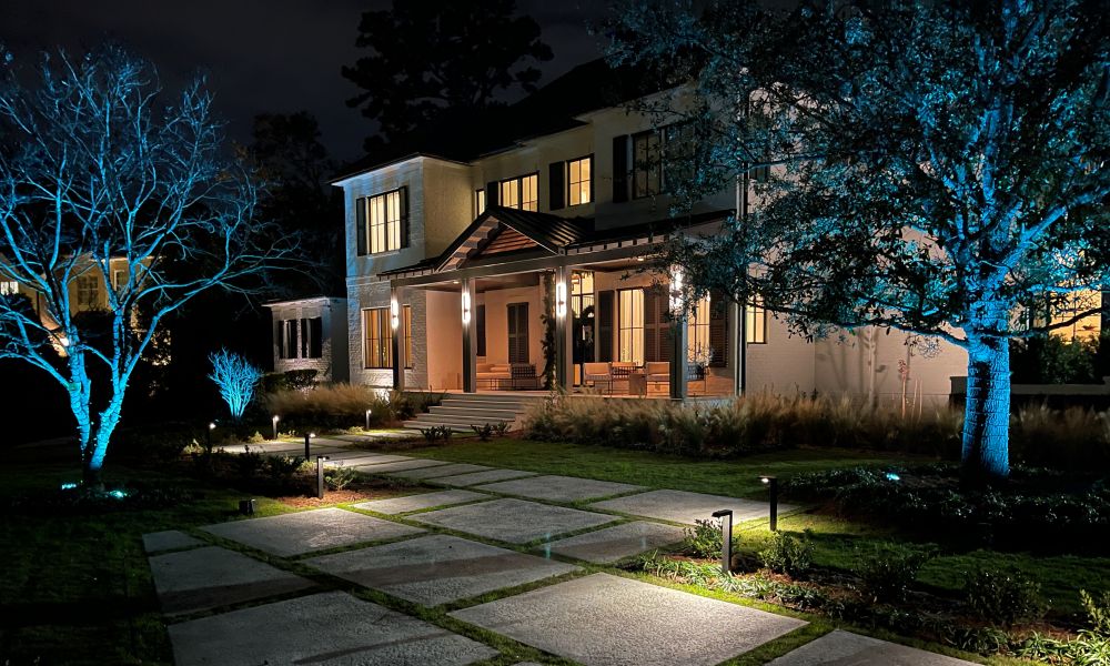 A two-story modern home is illuminated at night with pathway lights and blue accent lighting on surrounding trees.